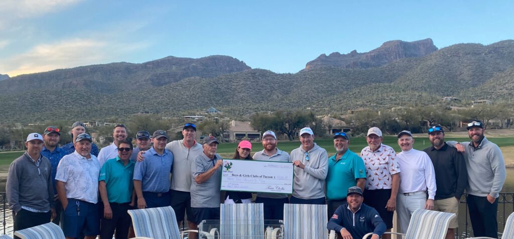 A large group of men and women are smiling for the camera and holding a large-format check. The golf course, mountains, and blue sky are in the background.